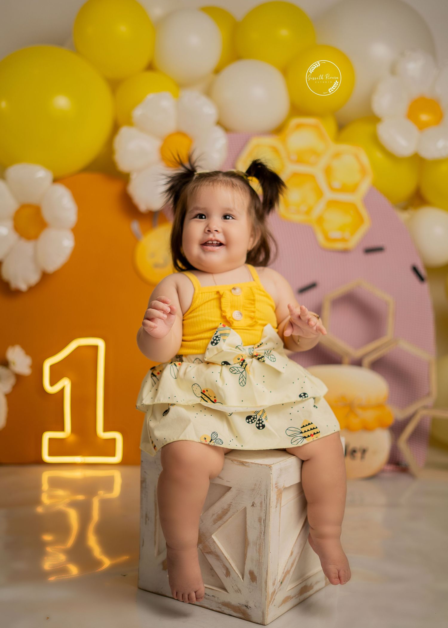 Happy baby girl in yellow bee outfit celebrates first birthday with balloon decoration backdrop.