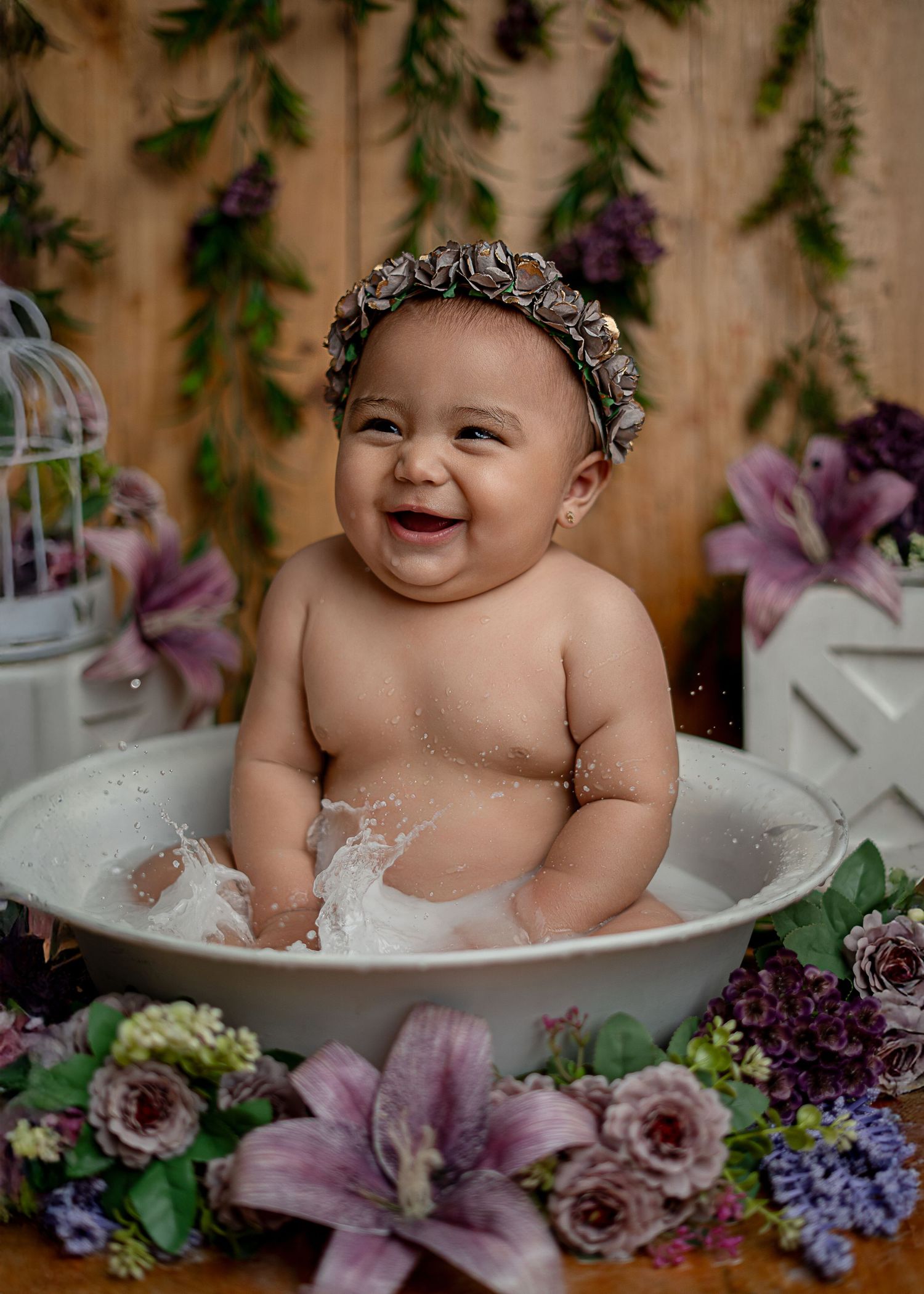 Cheerful baby with a floral crown laughing in a white tub surrounded by purple and pink flowers.