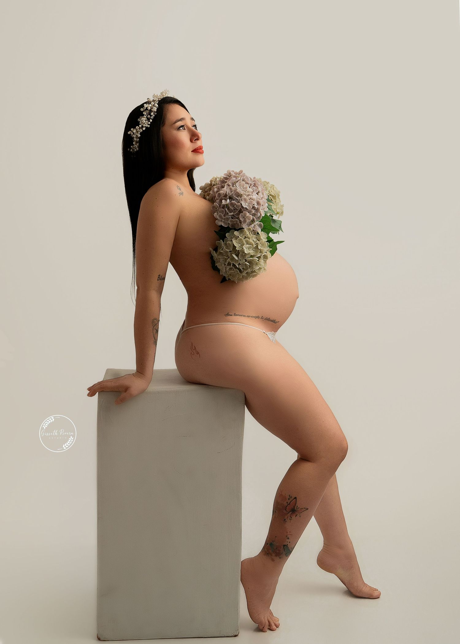 Pregnant woman in nude underwear poses sideways holding hydrangea flowers in minimalist studio setting.