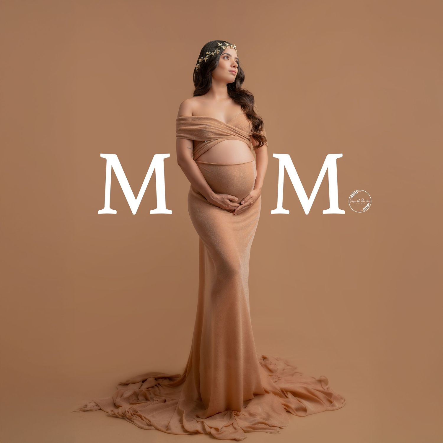 Pregnant woman in nude fitted maternity gown poses between decorative M letters in studio.