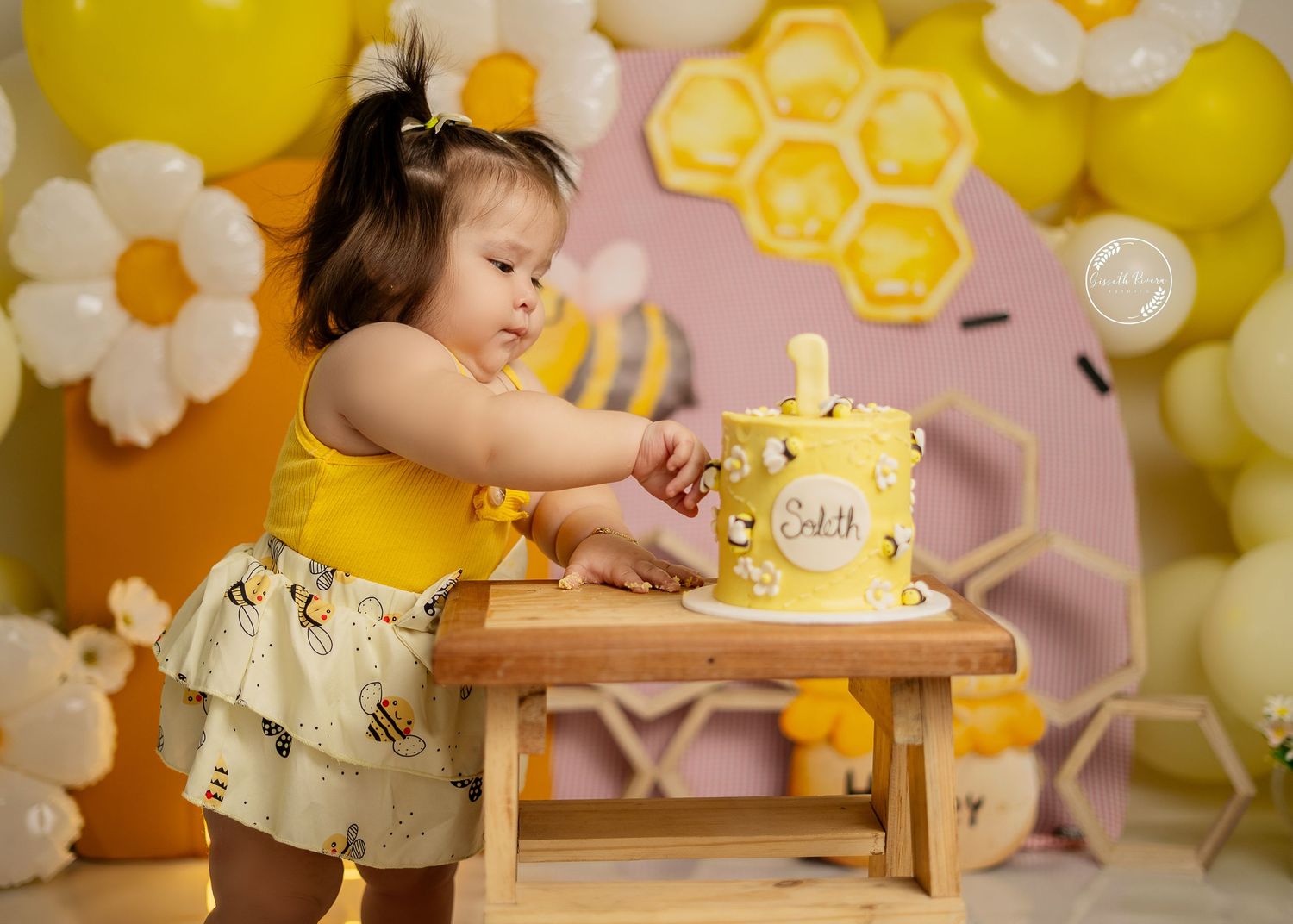 Baby girl in yellow outfit smashes first birthday bee-themed cake at decorated party setup.