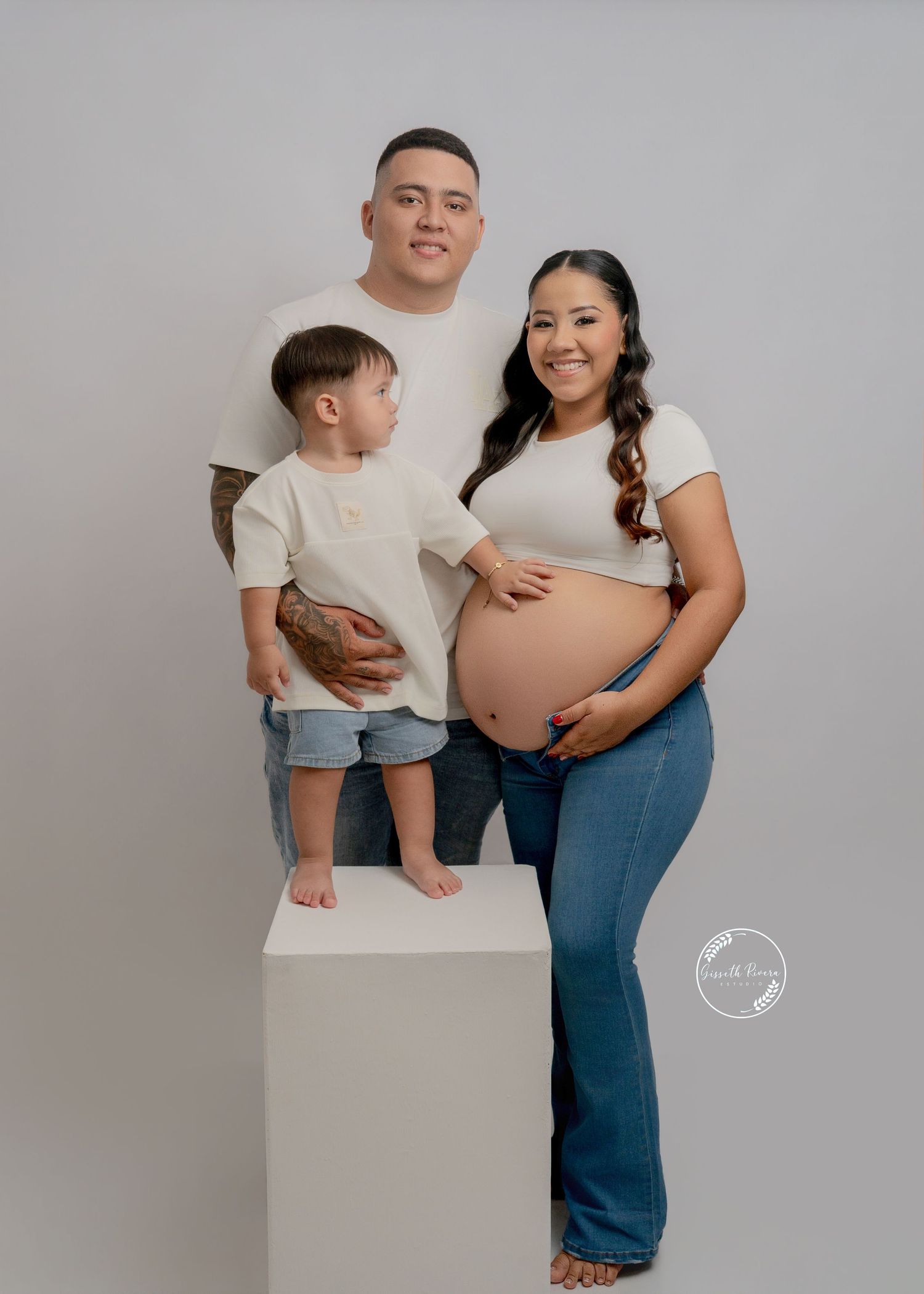 Family maternity photo with pregnant mother, father, and toddler in matching white outfits on gray backdrop.