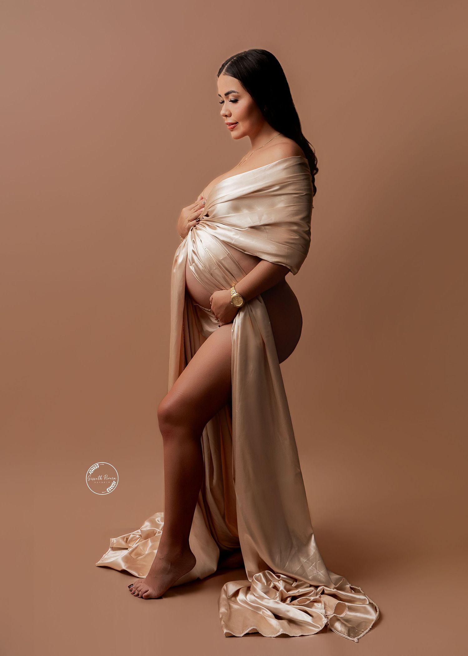 Elegant pregnant woman draped in golden satin fabric poses sideways in professional maternity portrait.