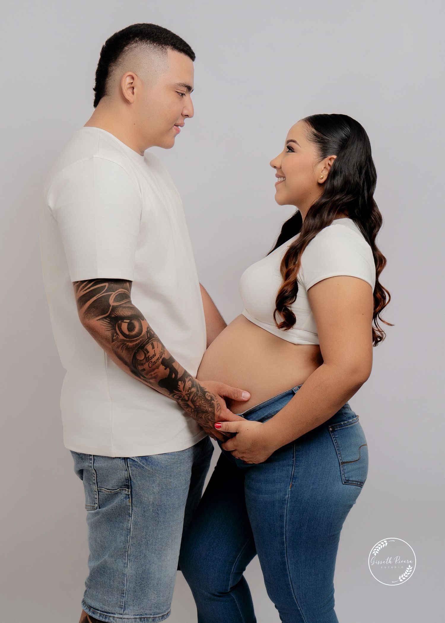 Couple maternity photo with tattooed man and pregnant woman in white outfits facing each other.