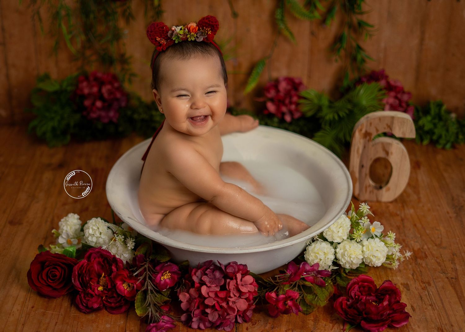 Laughing baby in a milk bath tub surrounded by red and white flowers on a rustic wooden floor.