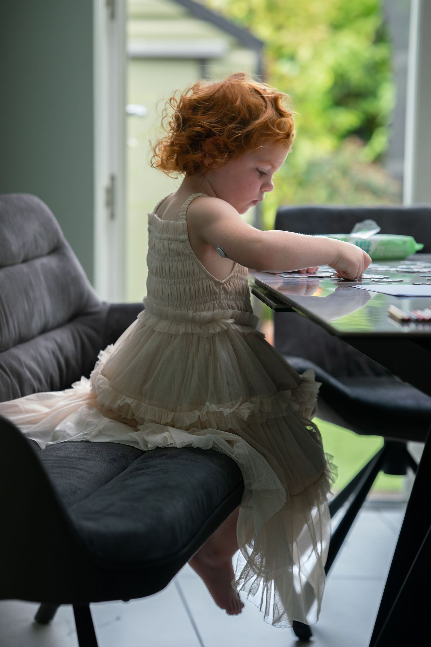 Young girl with curly red hair sitting on the sofa and doing a puzzle at the table, wearing a beige tulle dress at home.