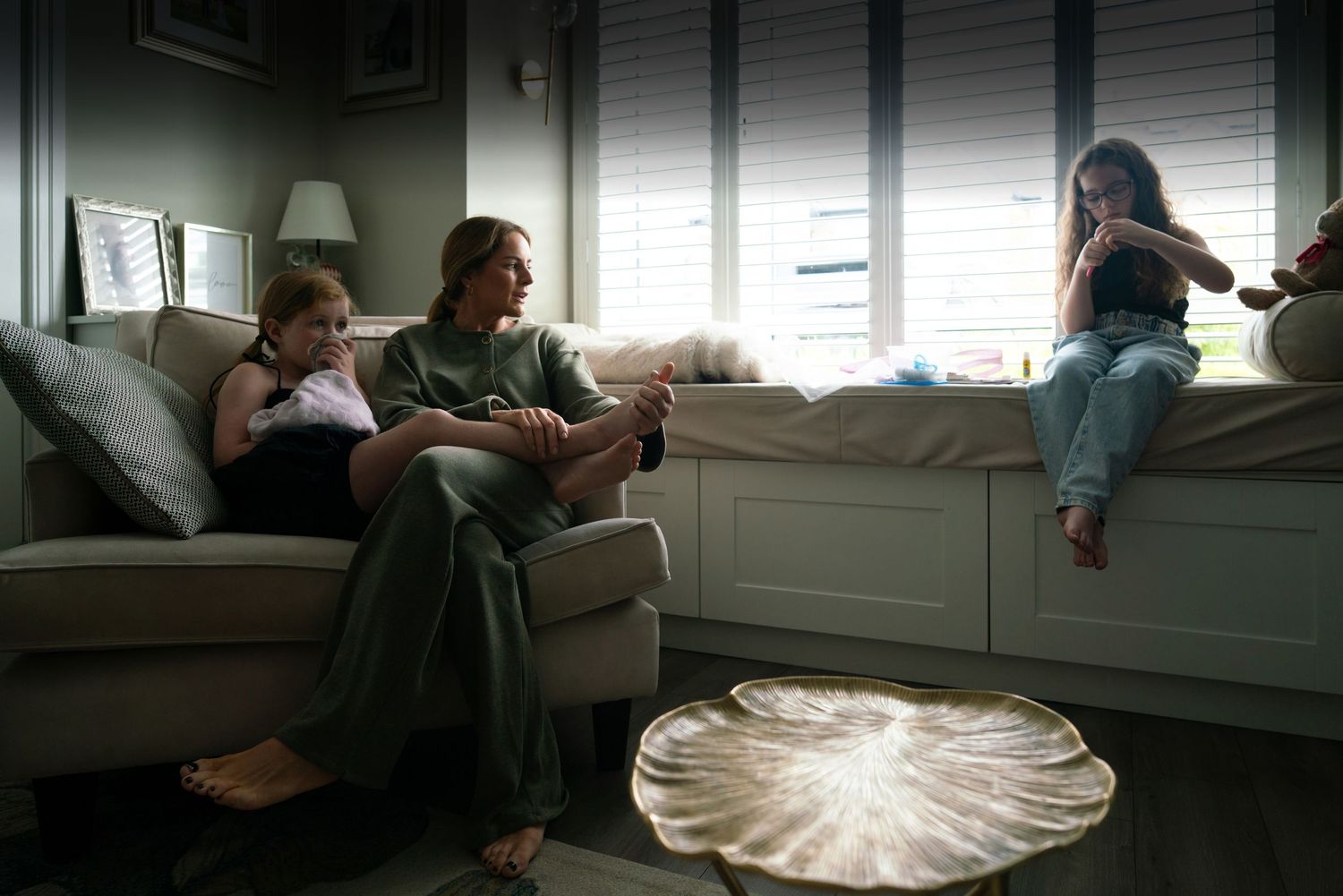 Mother relaxing with her daughter on the sofa while her other daughter does crafts by the bay window in their family living room.