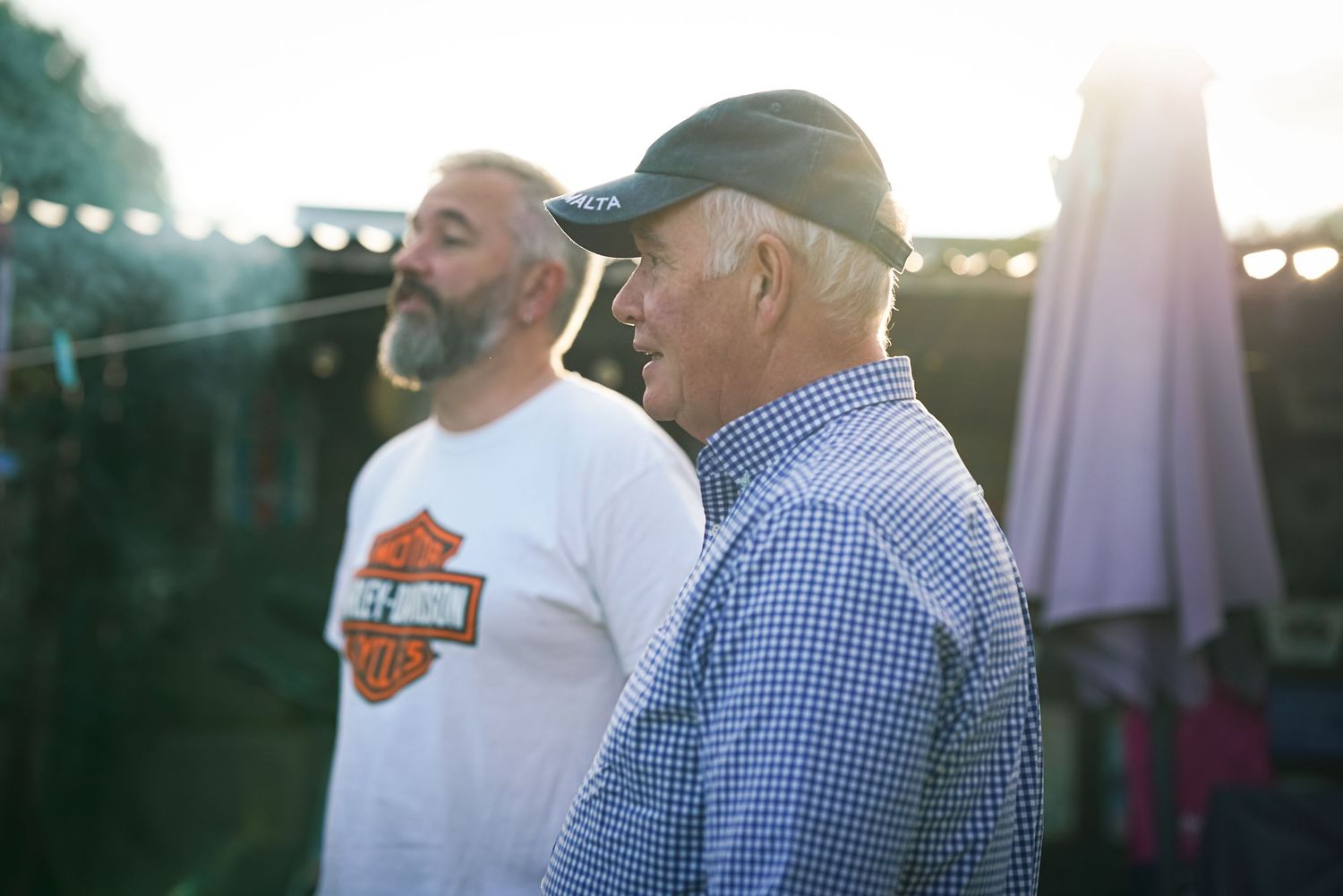Two men stand outdoors at a backlit evening gathering, one wearing a Harley-Davidson shirt.