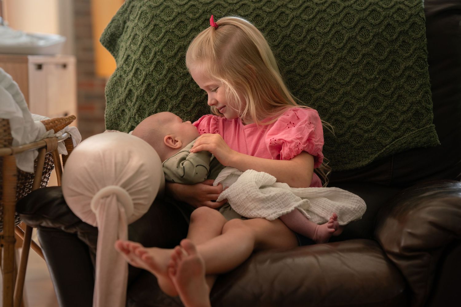 Young girl with long blonde hair smiling at her newborn brother as she holds him in her arms in a brown armchair at home.
