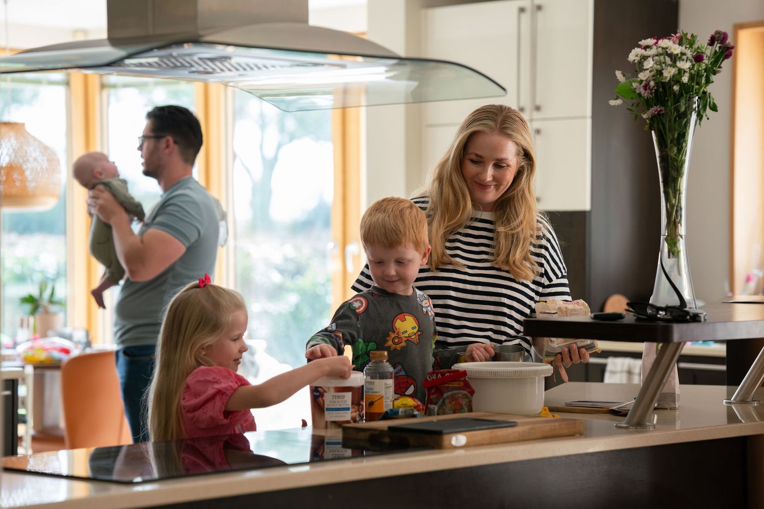 Mother and two children making cookies and chatting in their open-plan kitchen while their father holds their newborn baby in the background