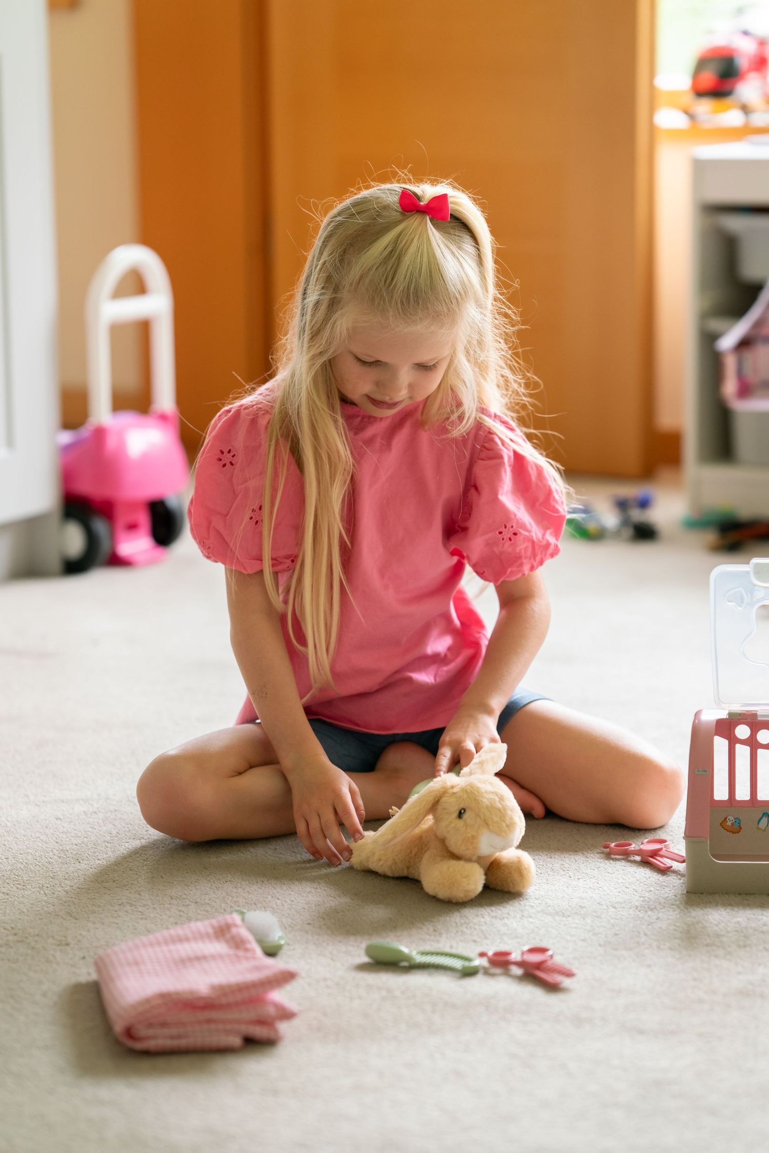 A blonde girl in pink plays doctor with a stuffed animal bunny on the floor.