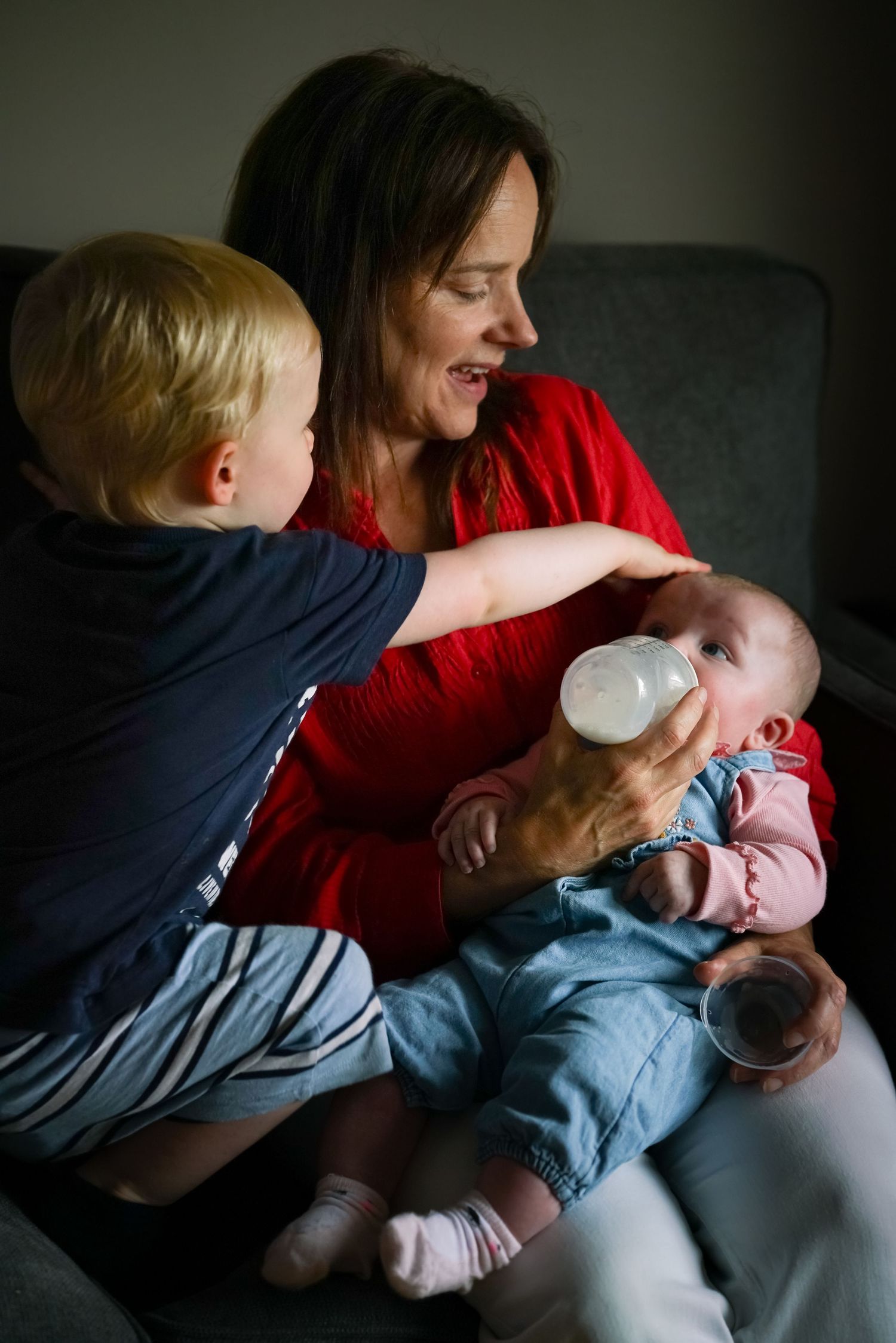 A smiling mother in red feeds a newborn with a bottle while a toddler watches.