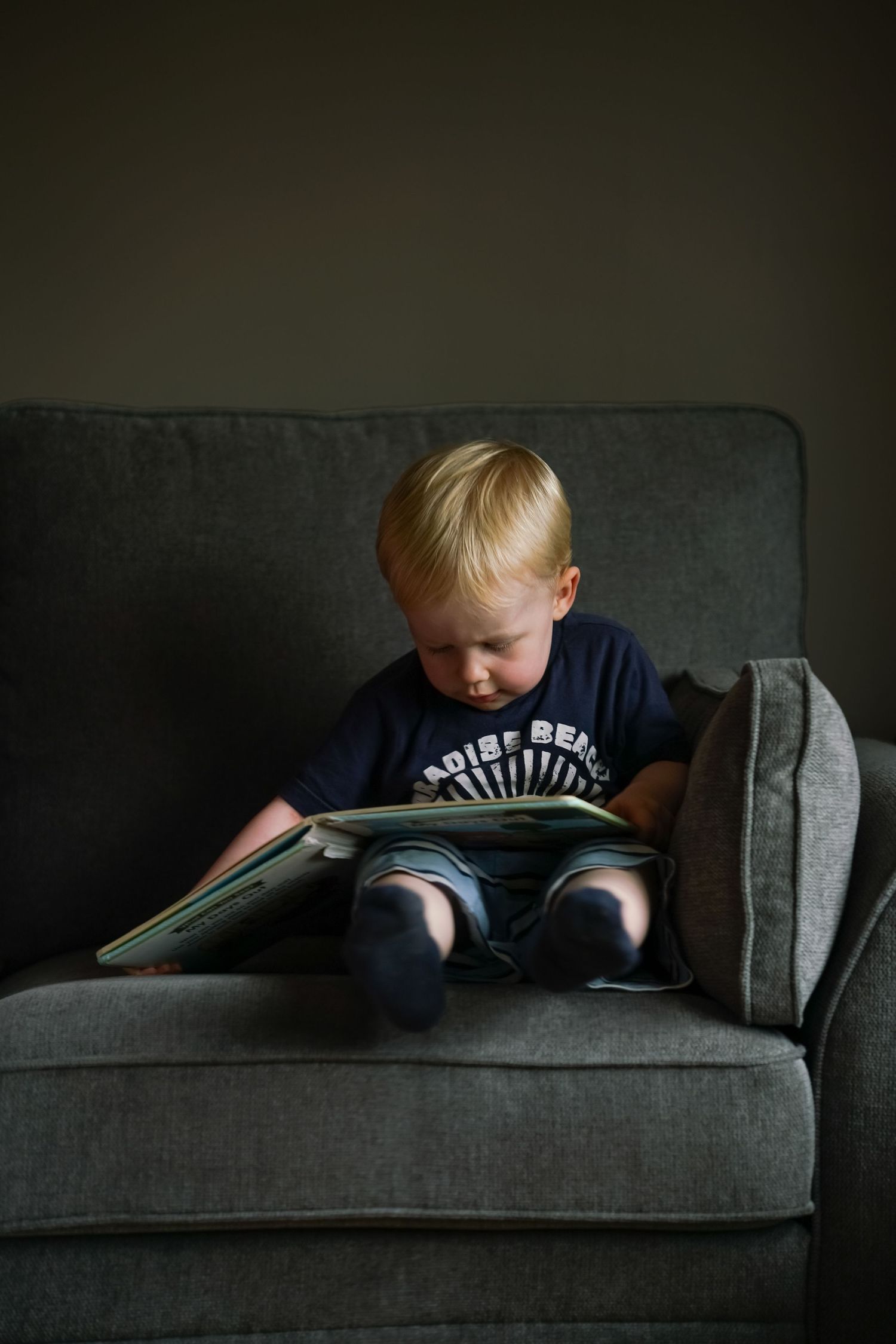 A blonde toddler sits alone on a grey sofa, focused on reading a picture book independently.