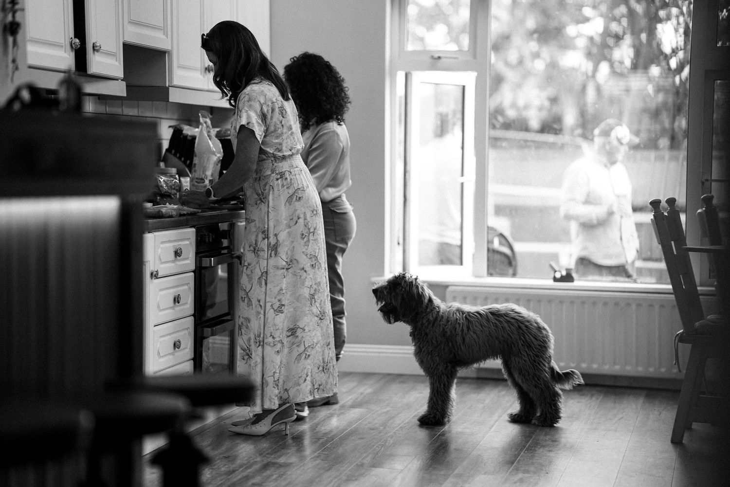 Two women stand in a kitchen while a shaggy dog watches them, in a black and white photo.