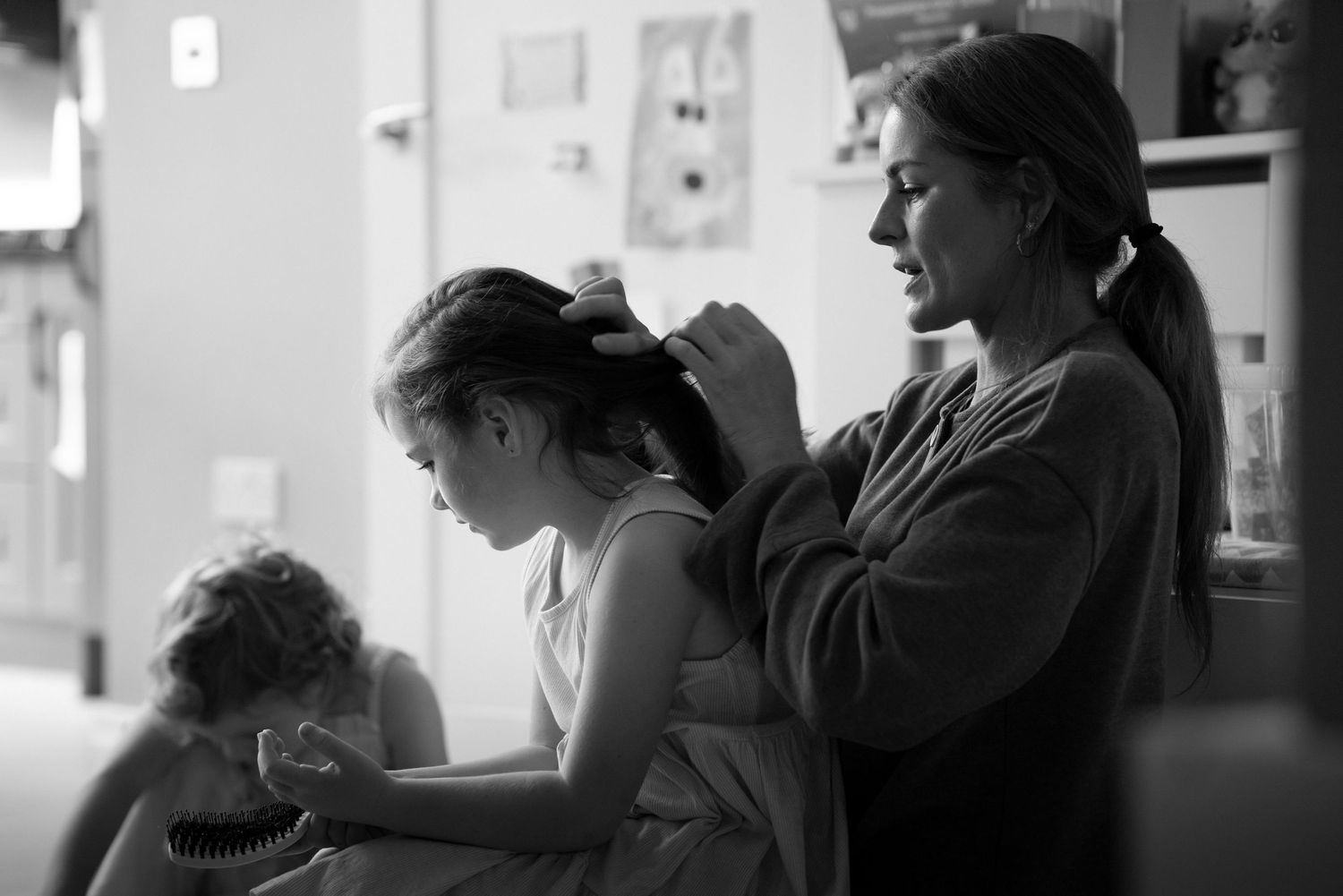 Mother tying her daughter’s hair at home while her other daughter plays in the background during an everyday family moment.