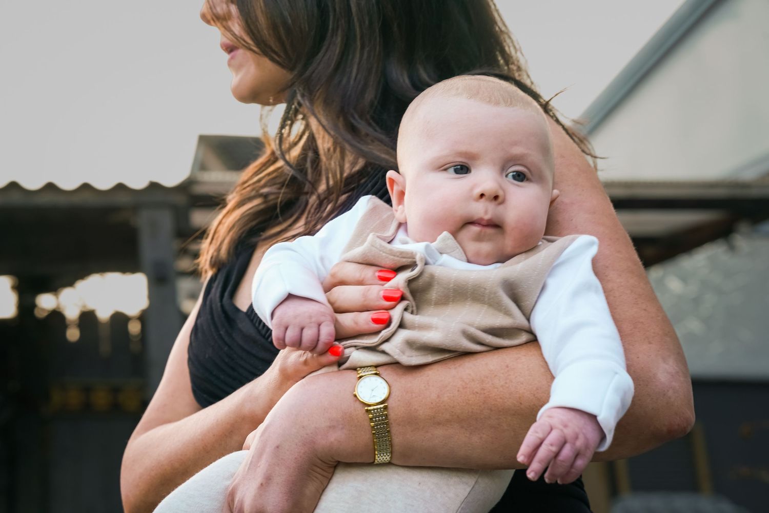 A woman with red nails holds an alert baby dressed in a beige outfit outdoors.