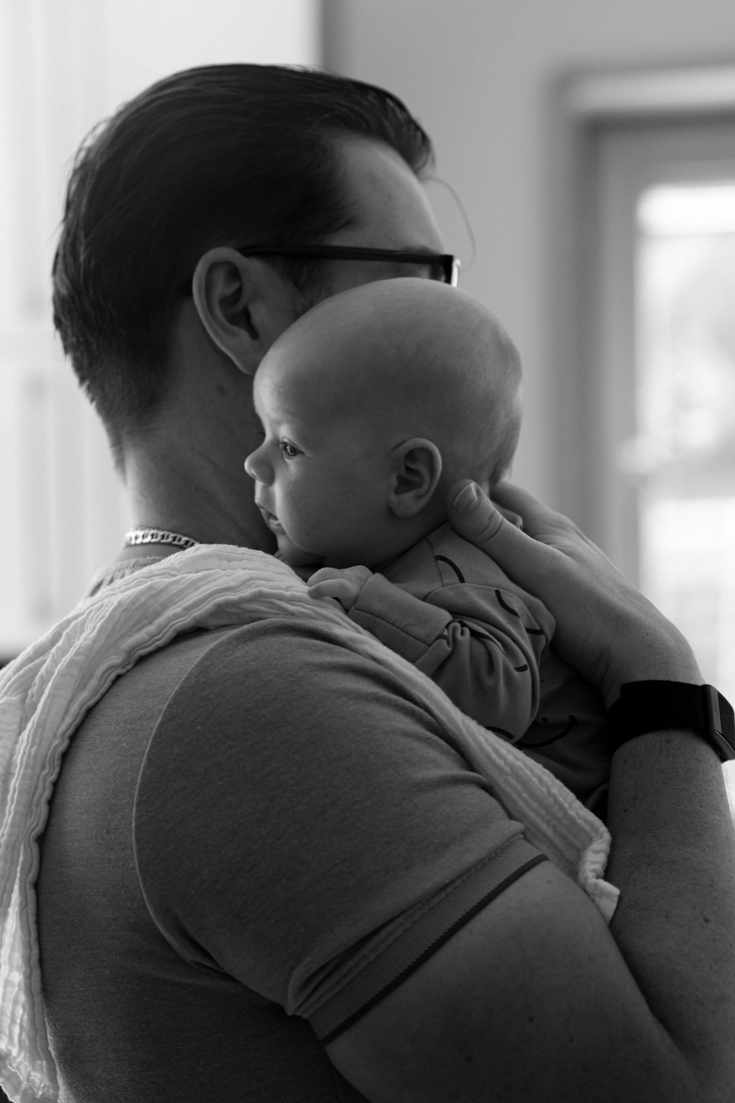 Black and white profile photograph of a father holding his newborn son against his shoulder, with a muslin cloth draped over him.