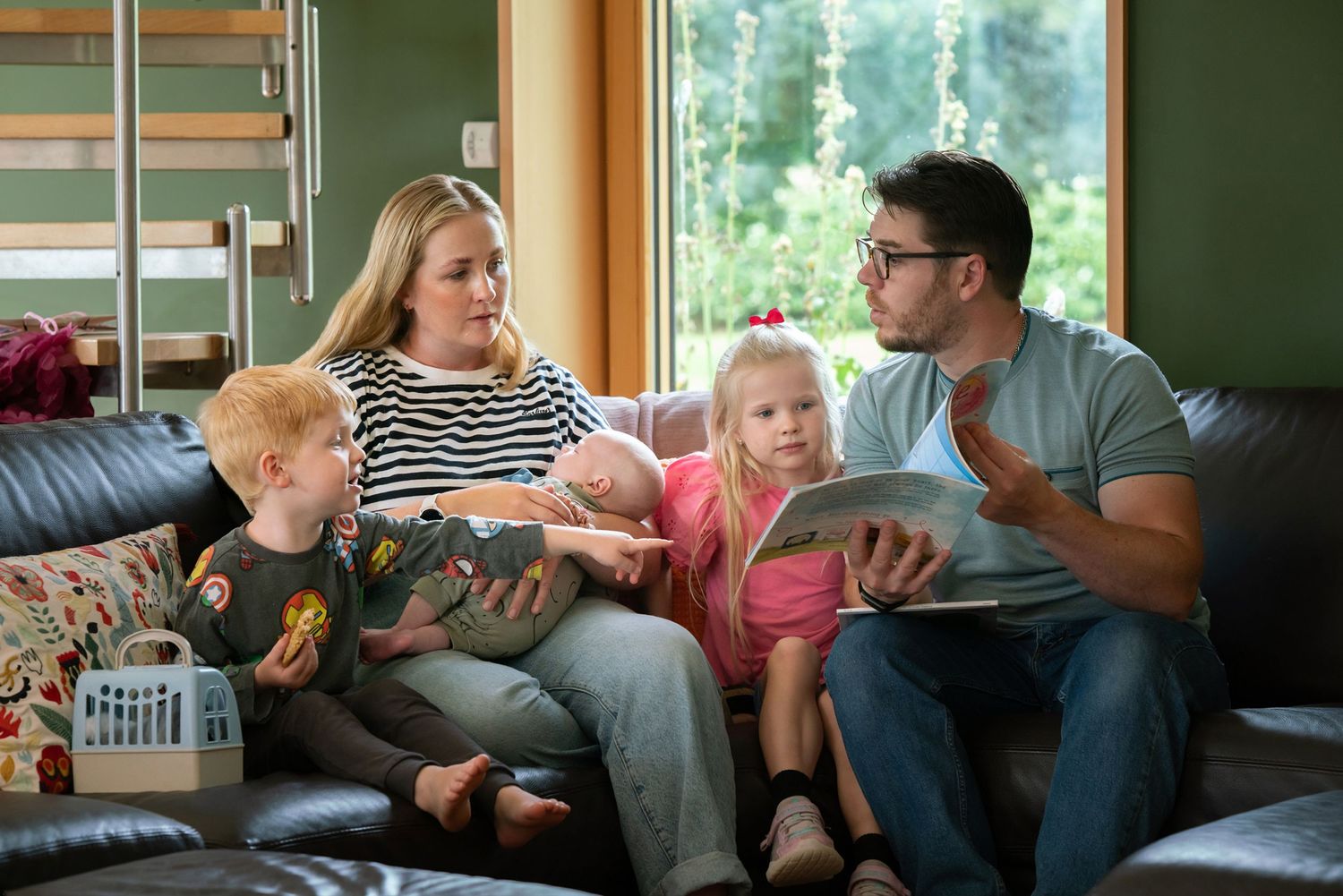 A young family with three children sits together on a leather sofa, reading a book in a cozy living room.