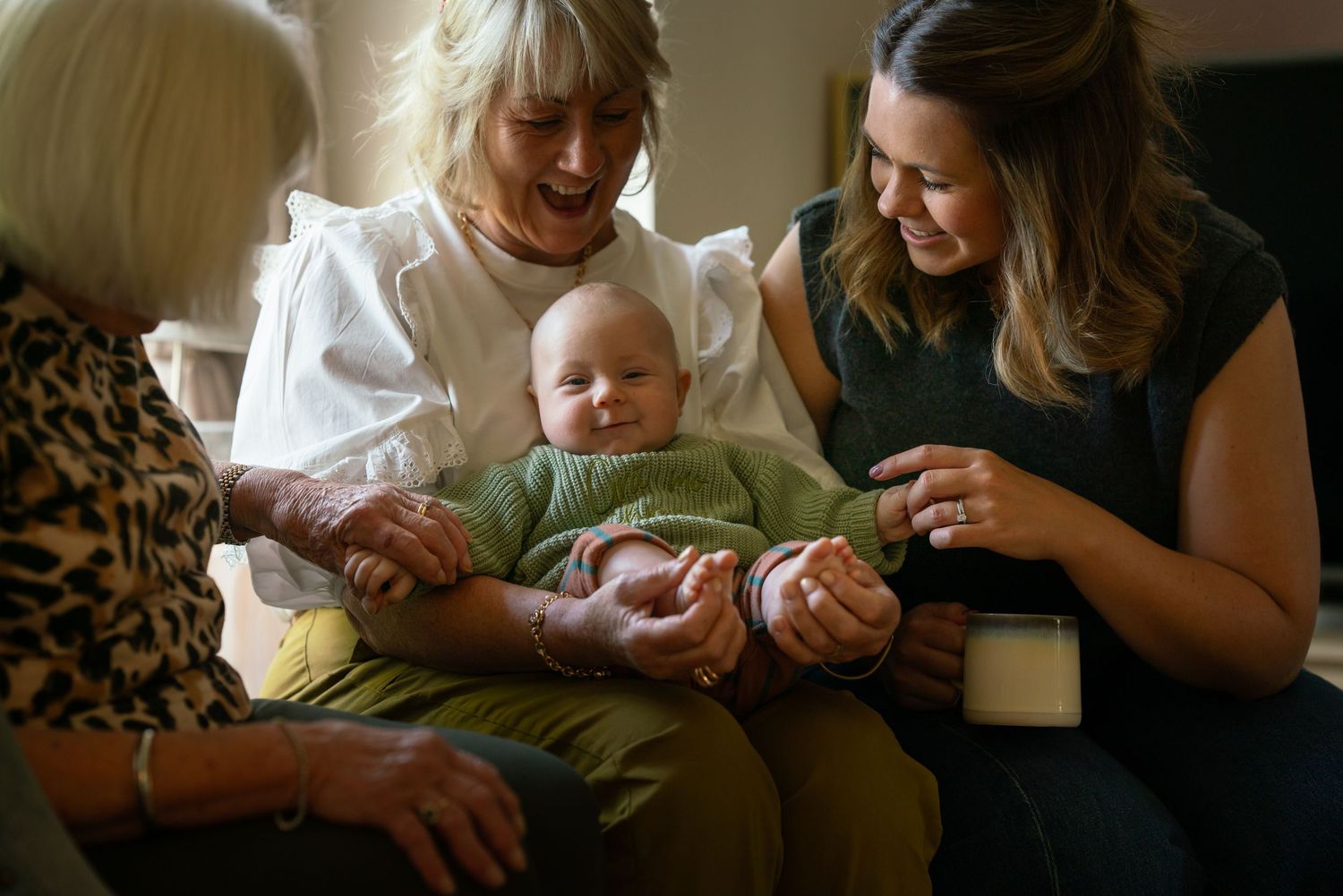 Three generations of women smile while holding a newborn baby indoors.