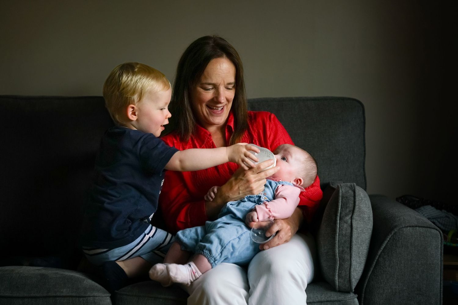 Family relaxing together on the sofa as a toddler gives his newborn sister a bottle while their mother holds her, smiling beside them.