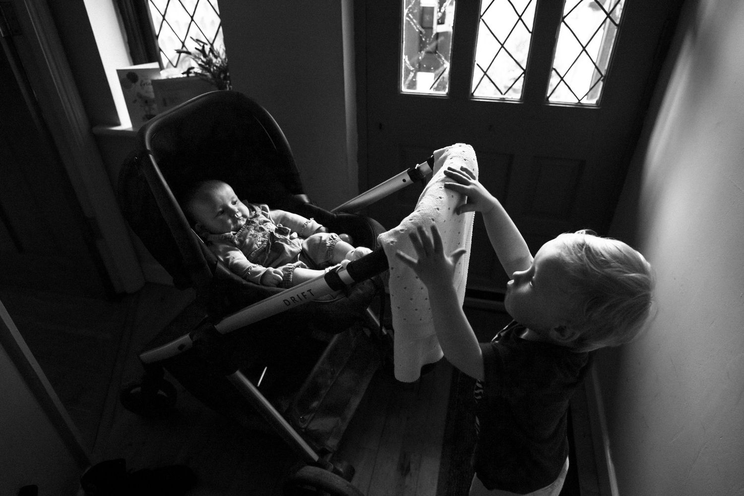 Black and white photograph of a toddler pushing his baby sister in her pram through the hallway at home.