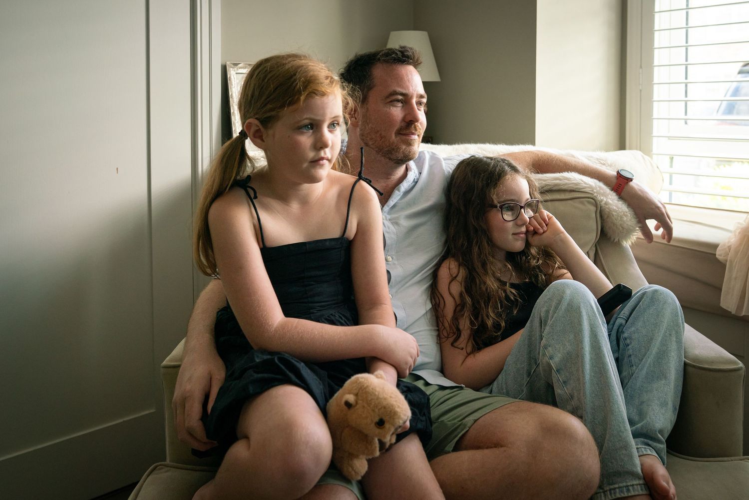 A father sits with his two young daughters on a couch near a window.