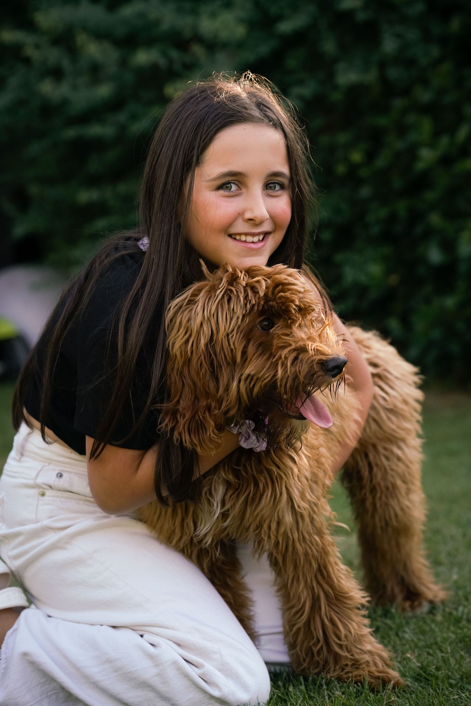 A smiling girl hugs her fluffy golden Labradoodle dog on green grass outdoors.