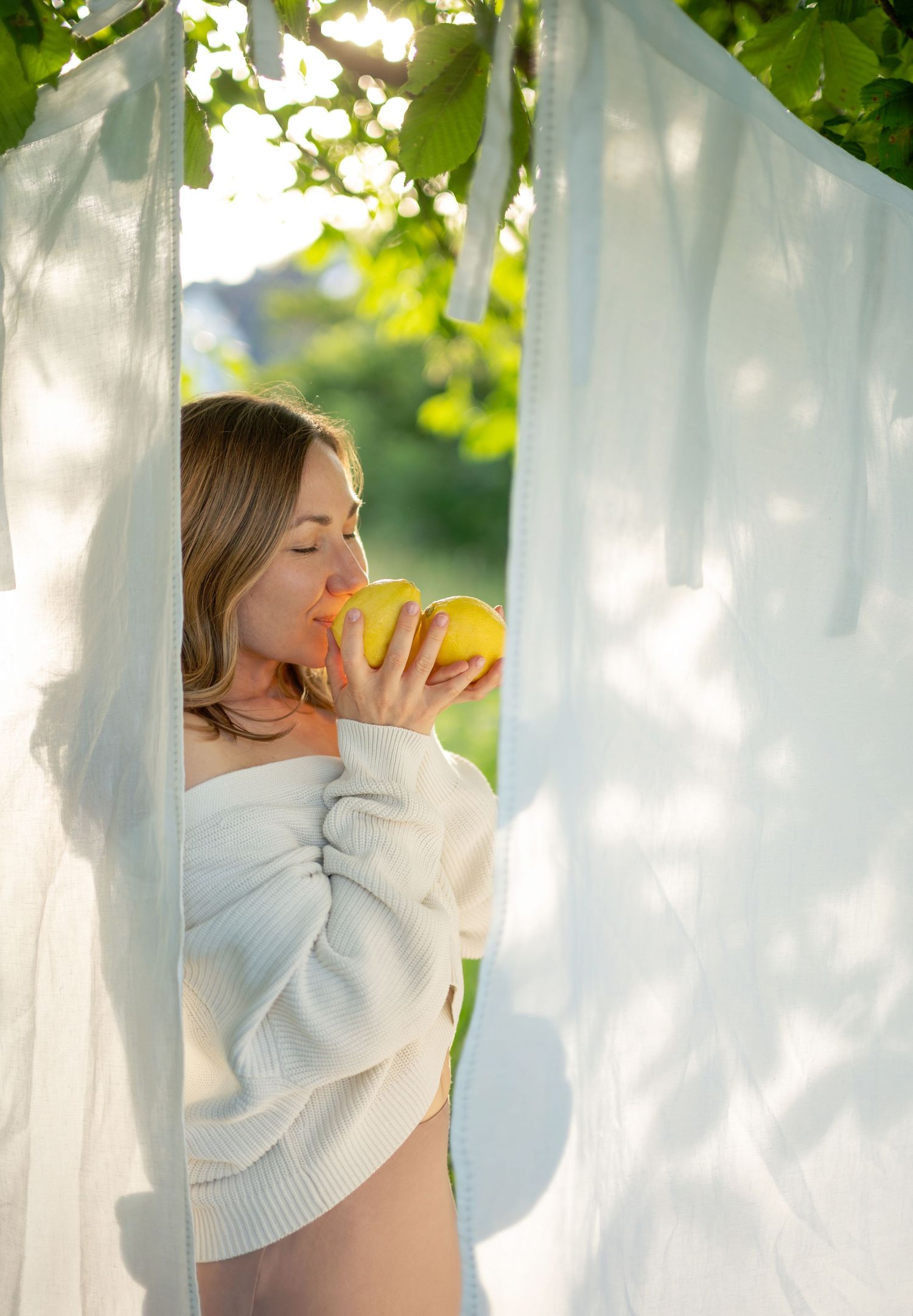 Pregnant woman in white off-shoulder sweater smelling a lemon outdoors, framed by sheer white curtains and green foliage.