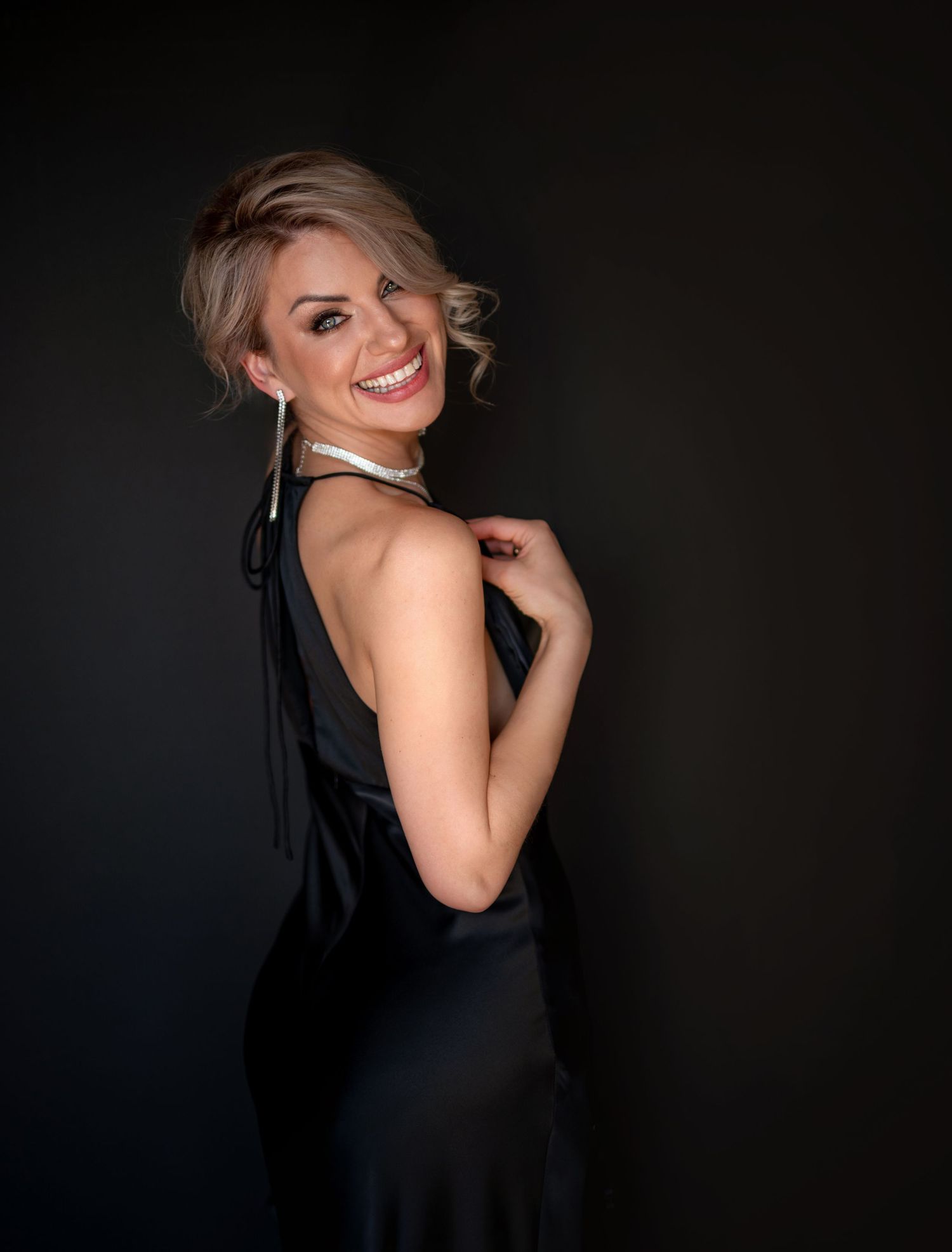 Smiling blonde woman in elegant black backless gown with pearl necklace and earrings posing against dark background.