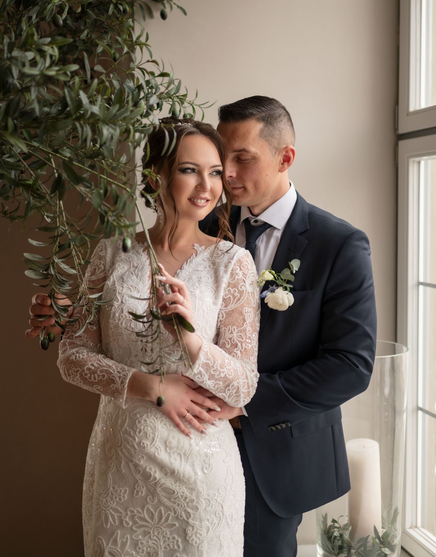 Bride in lace gown and groom in navy suit embrace near window with greenery at elegant wedding ceremony.
