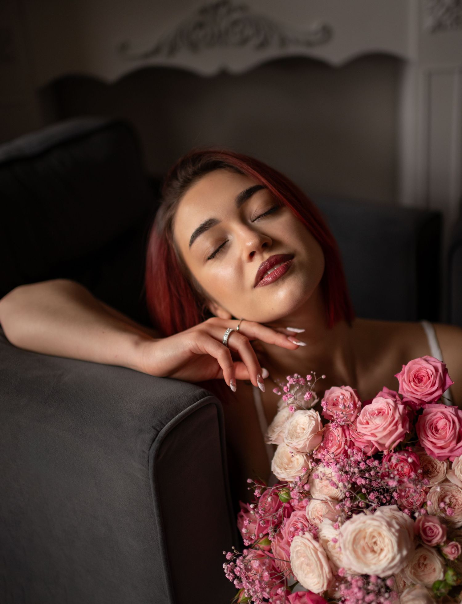 A young woman with red hair relaxes on a gray sofa, eyes closed, holding a bouquet of pink roses.