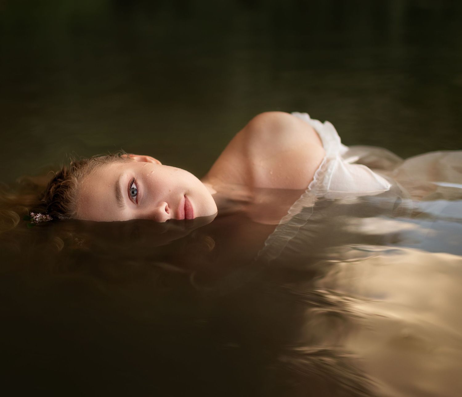 Young woman in white dress floating in dark reflective water looking at camera.