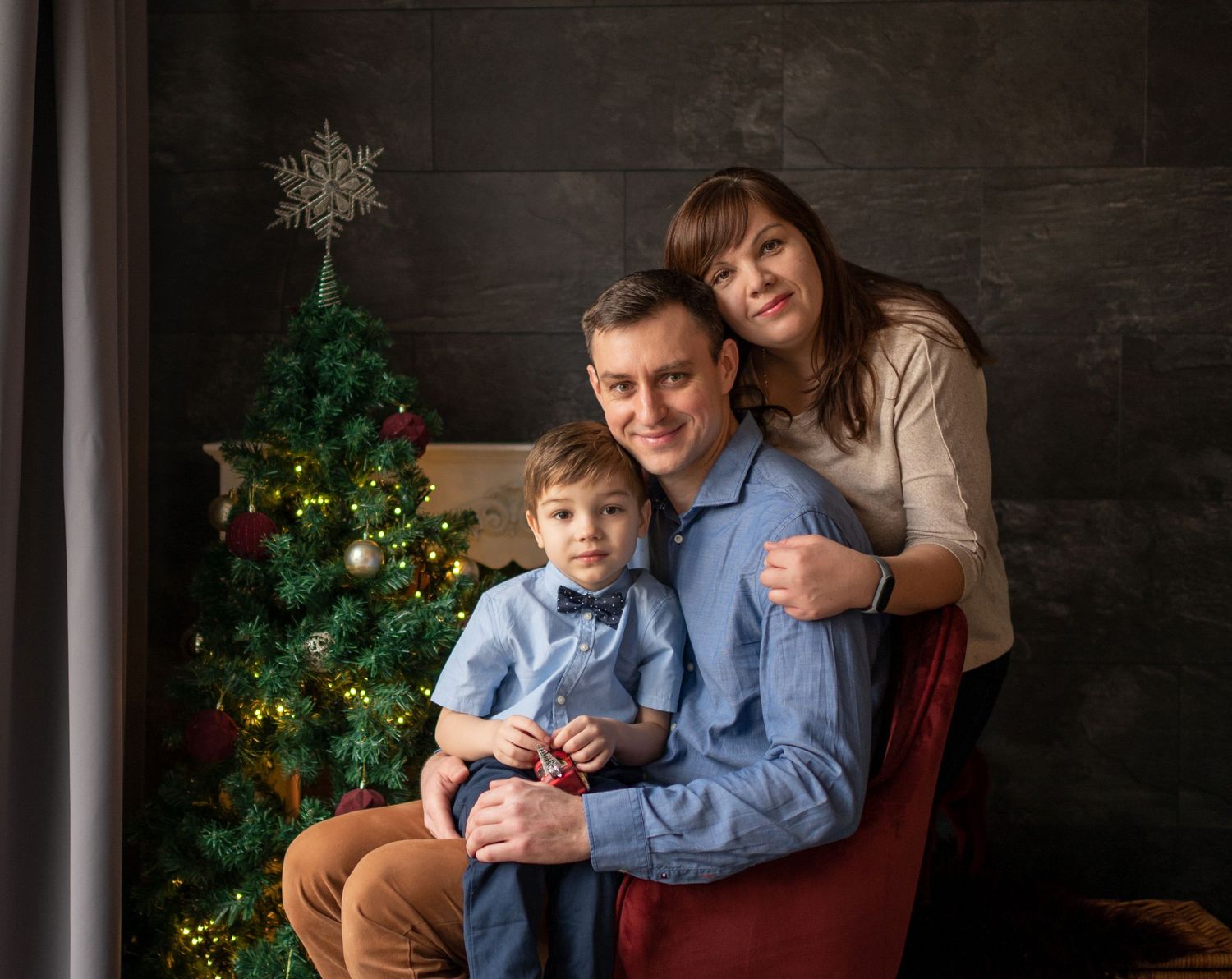 Weihnachtliches Familienfoto in Ulm Eltern küssen sich vor einem geschmückten Weihnachtsbaum, während ihr Kind eine goldene Geschenkbox hält