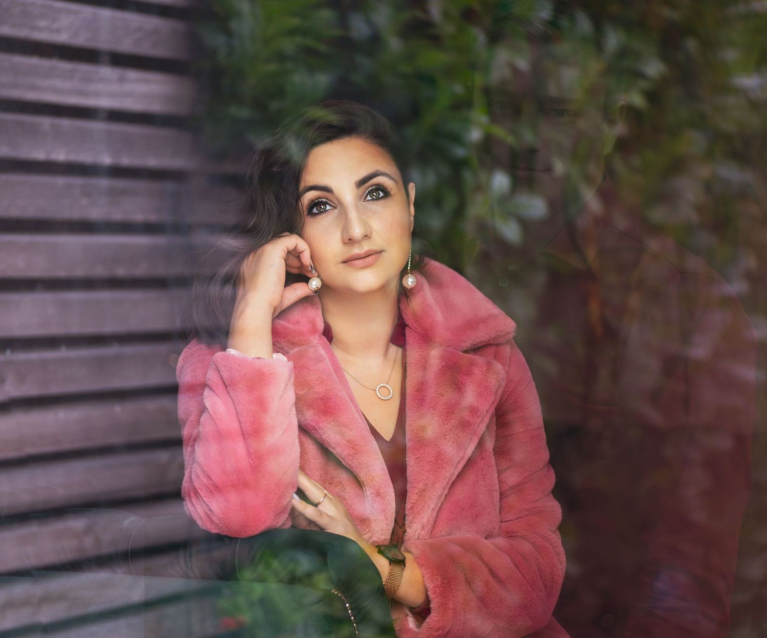 Young woman in pink faux fur coat posing thoughtfully against a wooden wall with green foliage backdrop.