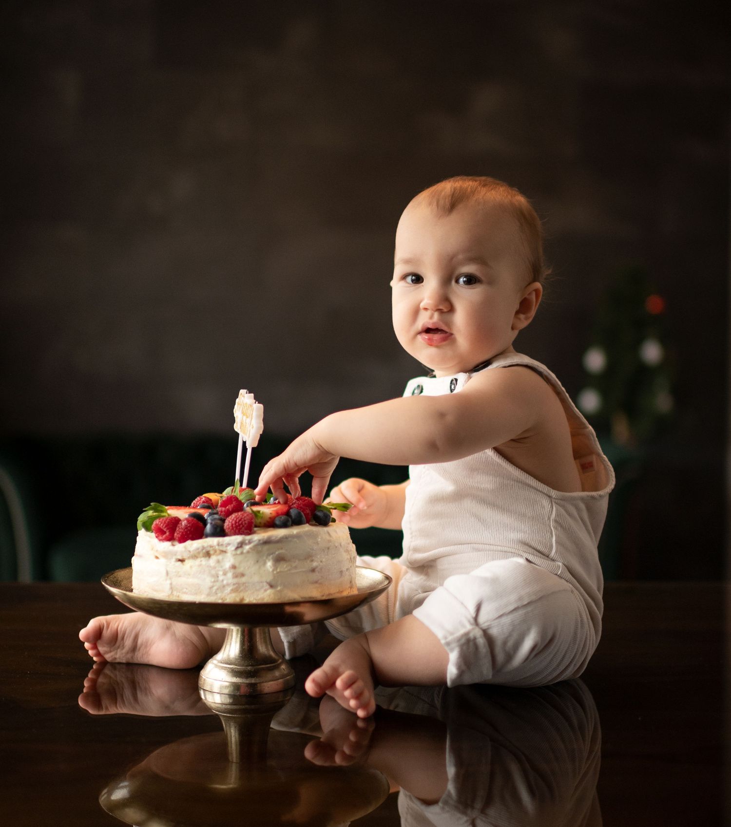 Adorable baby in white romper reaches for berry-topped cake on dark wooden table.