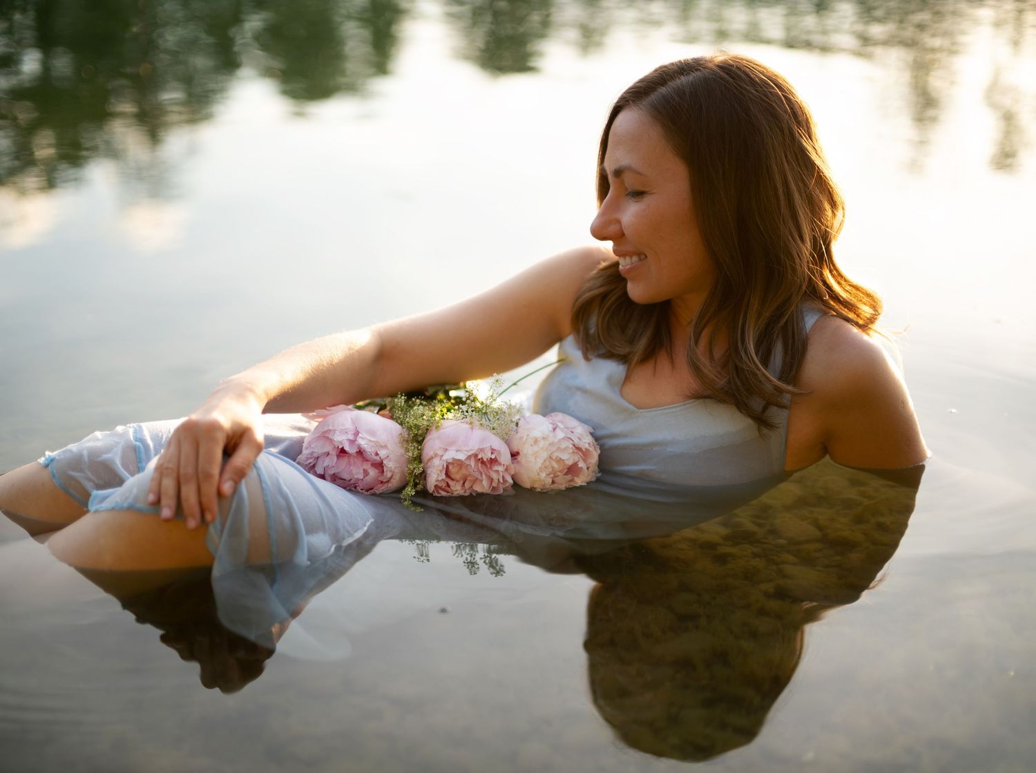 Woman in white dress holding pink peonies while lying in calm reflective water during golden hour.