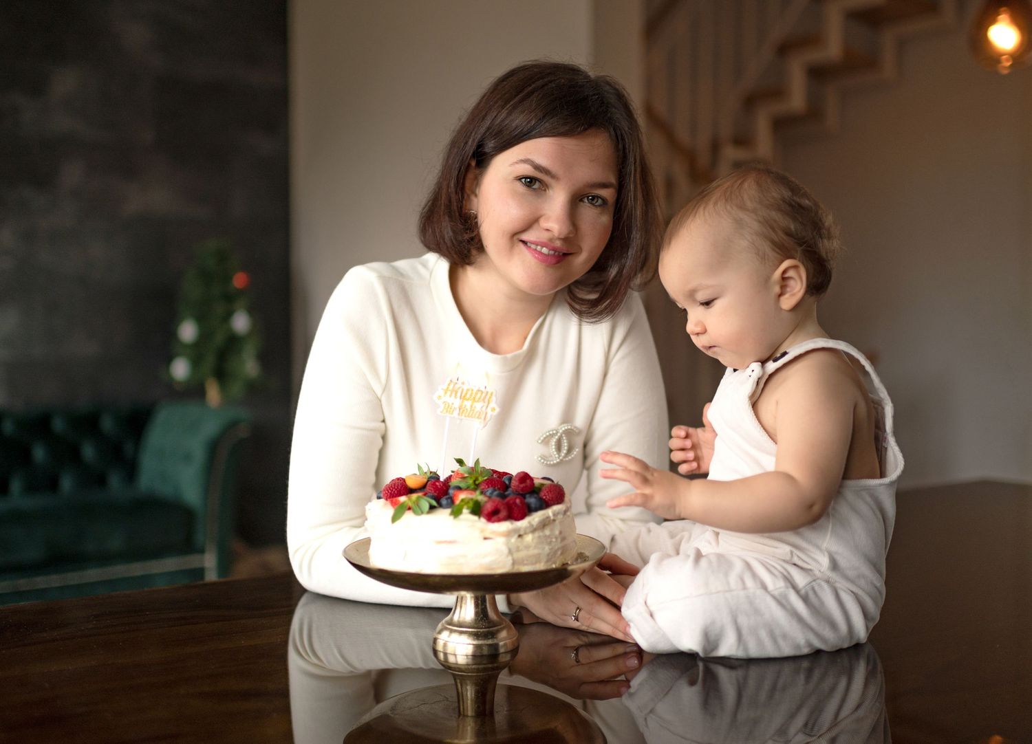 Young mother holding baby near birthday cake with fresh strawberries indoors.
