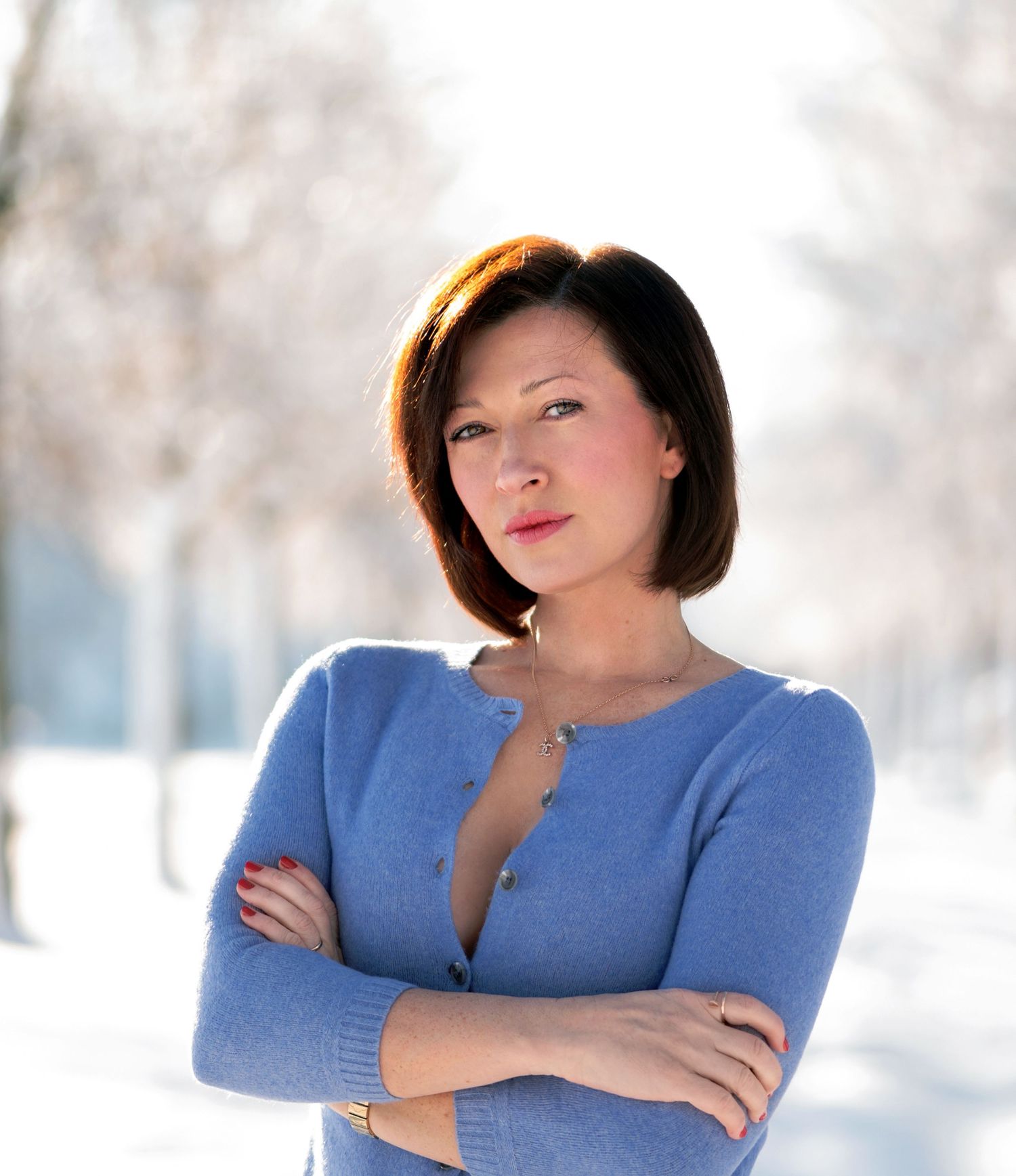Woman in blue cardigan with arms crossed standing outdoors in snowy winter park setting.
