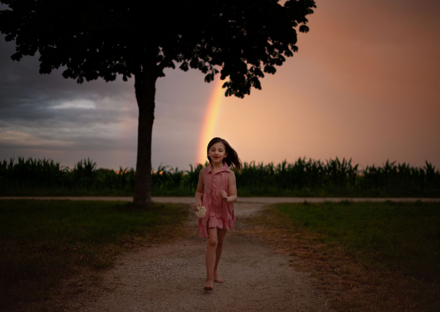 Young girl in pink dress walking on path with rainbow in stormy sky.