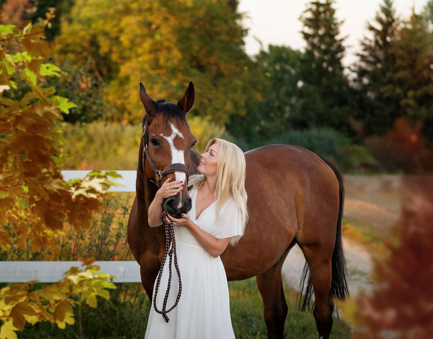 Blonde woman in white dress affectionately nuzzling brown horse in autumn setting.
