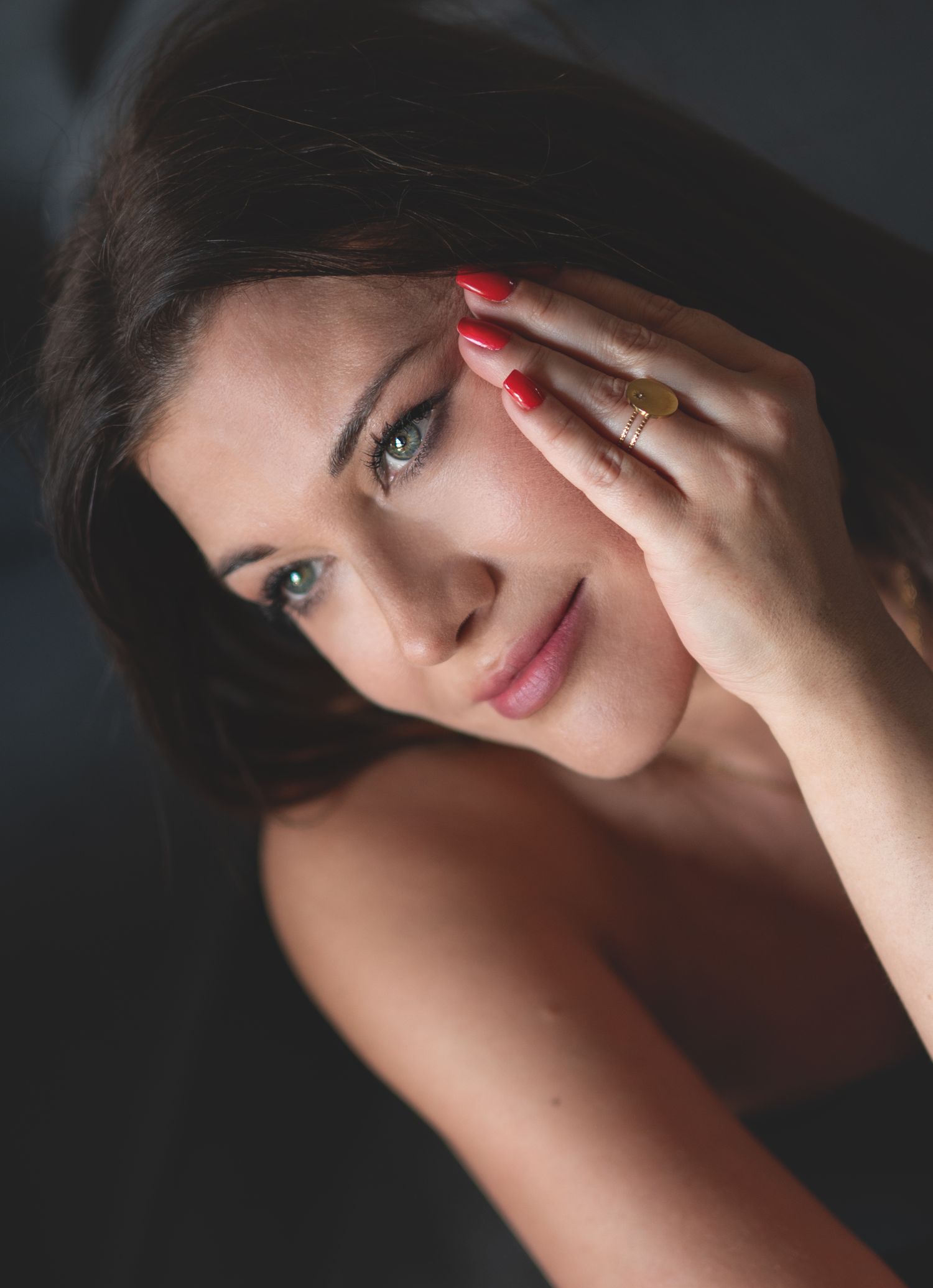 A young woman with dark hair and red nails rests her hand near her face, wearing a gold ring, in a moody portrait.