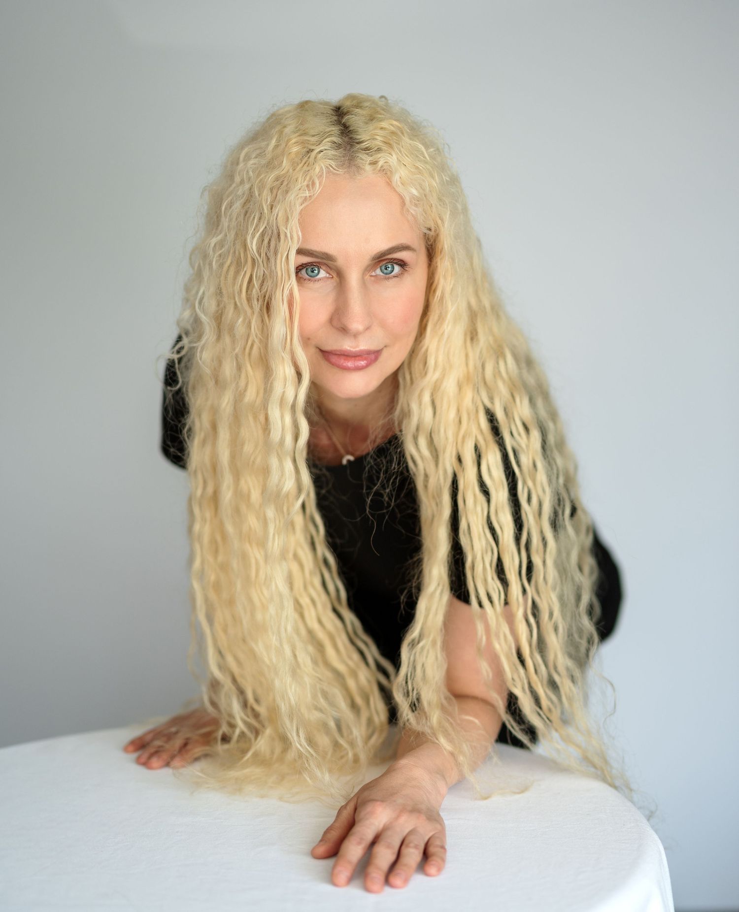 Woman with long, curly blonde hair wearing a black top, posing with arms on a white table against a gray background.