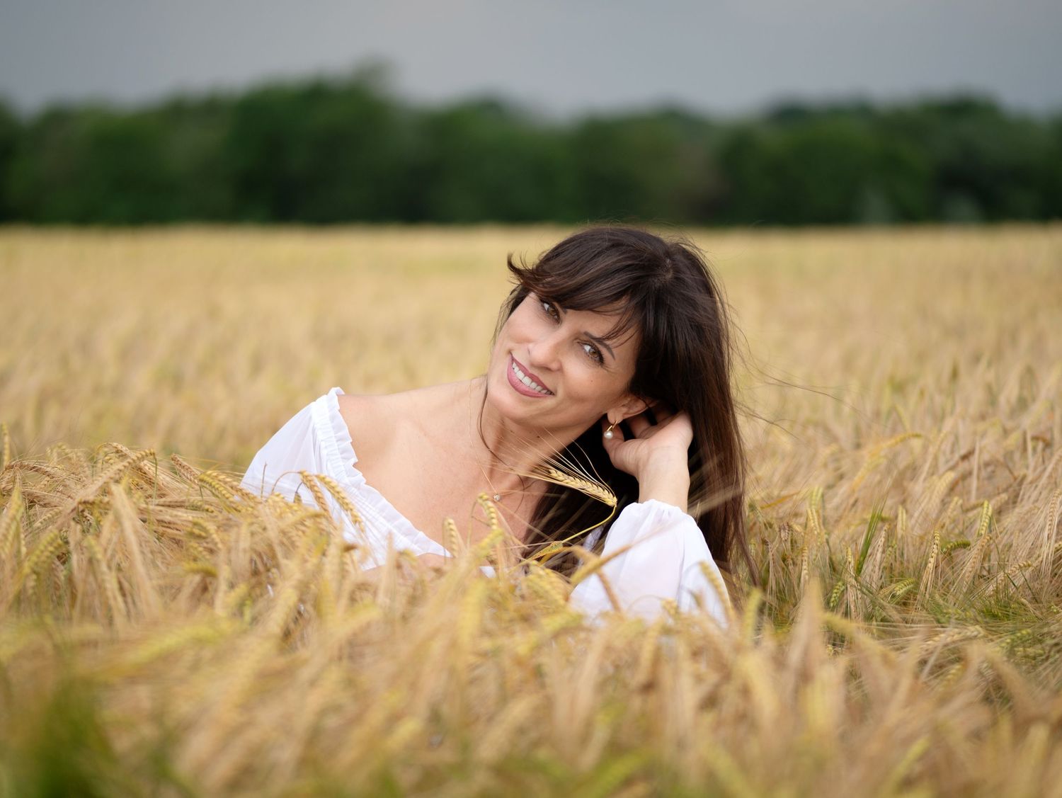 A smiling brunette woman in a white dress relaxing in a golden wheat field with green trees in the background.