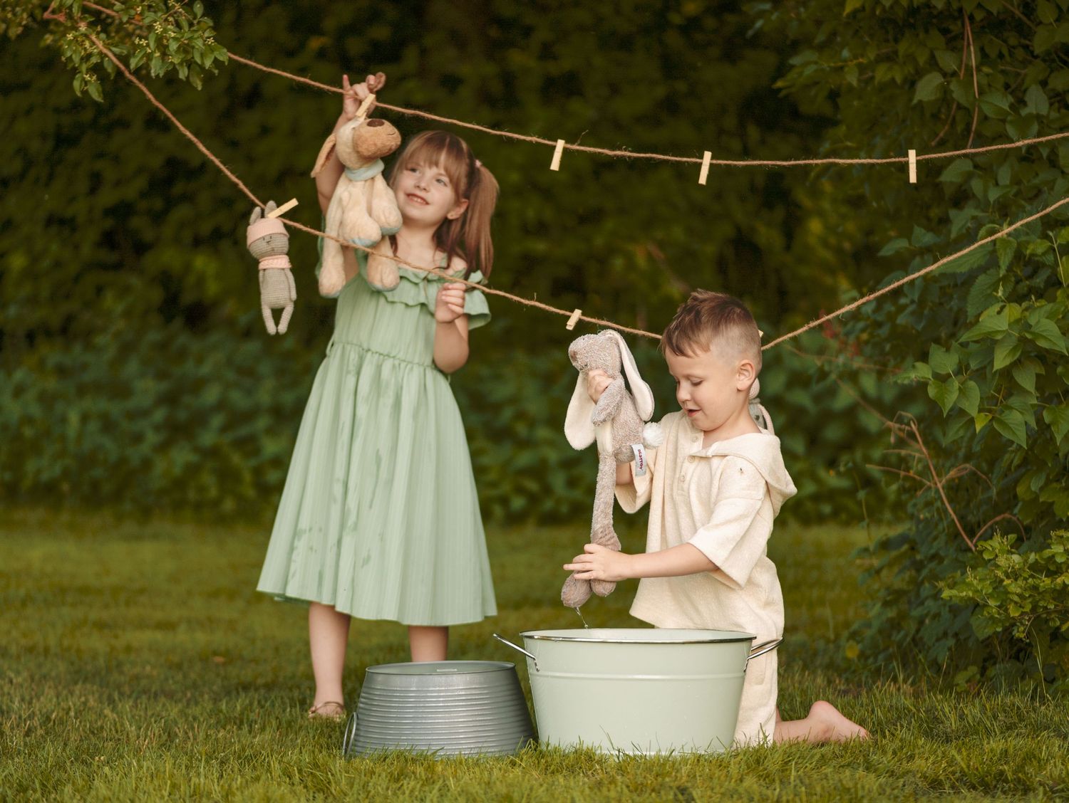 Two children play outdoors washing stuffed animals in metal tubs near a clothesline.