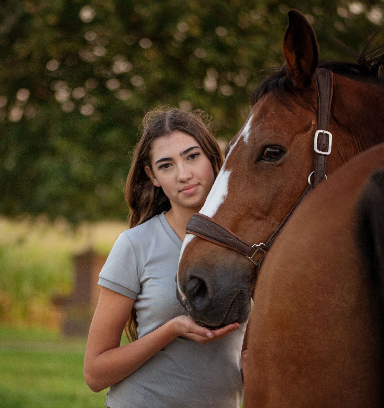 Young woman with brown hair standing beside brown horse in outdoor setting.