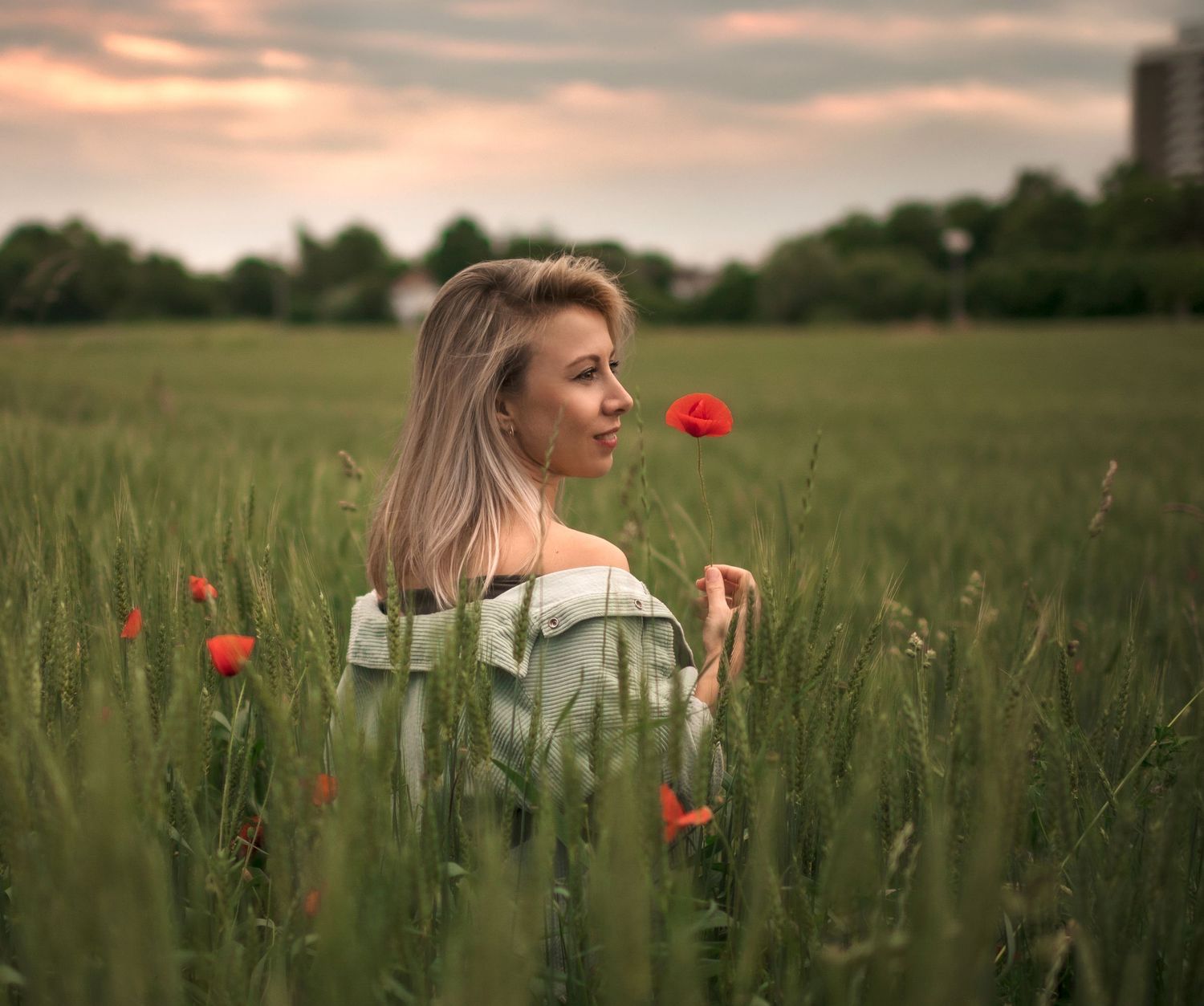 Young blonde woman holding a red poppy flower in a lush green field at dusk.