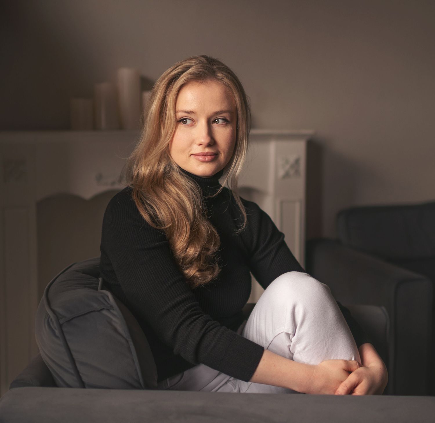 Young woman with long blonde hair wearing black turtleneck and white pants sitting on a couch indoors.