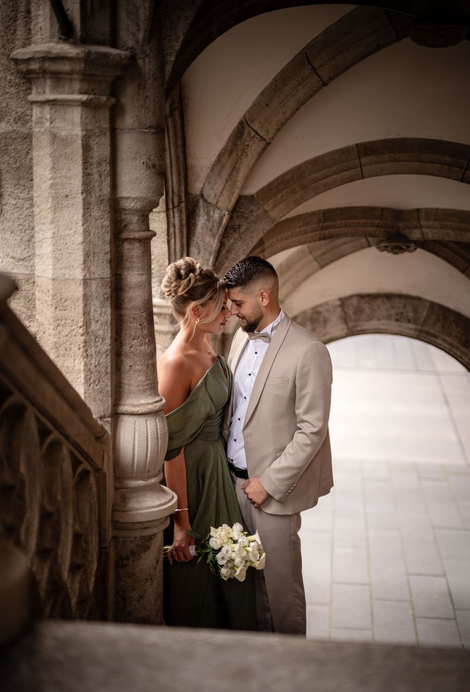 Elegant couple in formal attire sharing a romantic moment under stone arches, woman in green gown holding white flowers.
