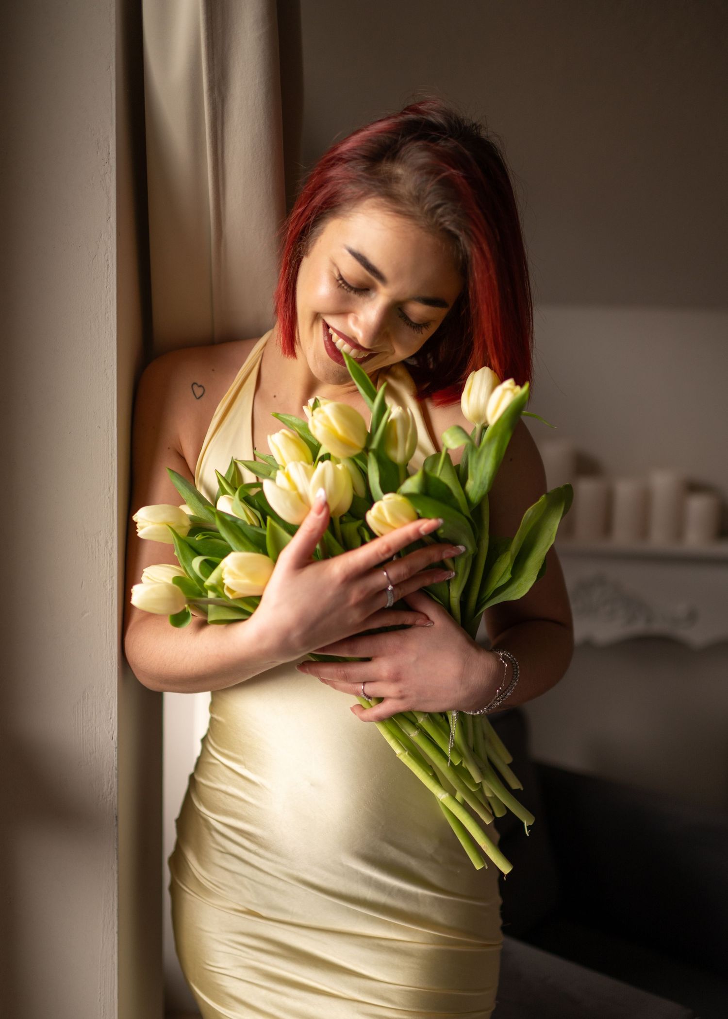 Smiling woman with red hair in gold dress holding white tulips bouquet near window.
