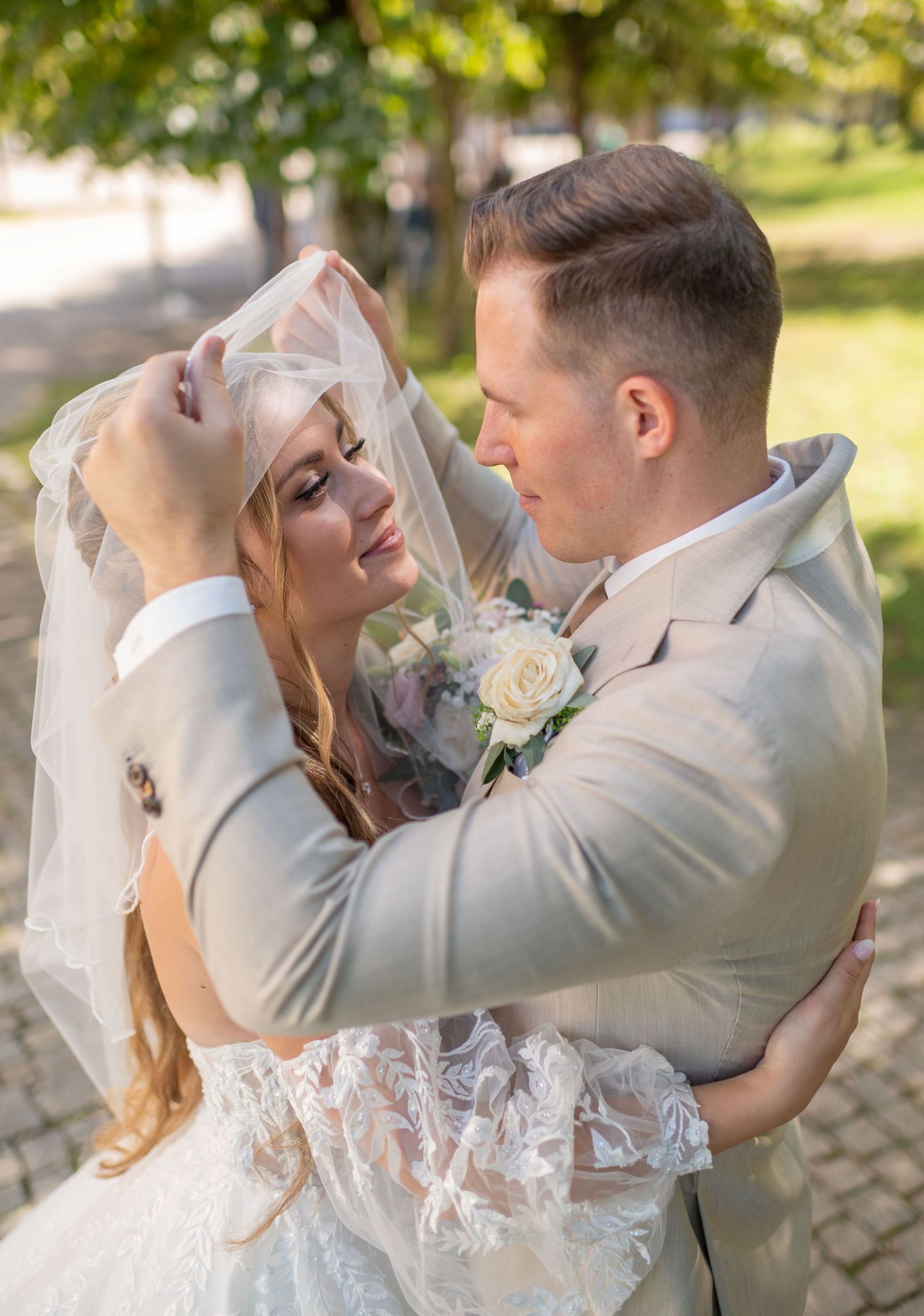 Bride and groom share a romantic moment under veil outdoors, groom in gray suit with white boutonniere, bride in lace gown.